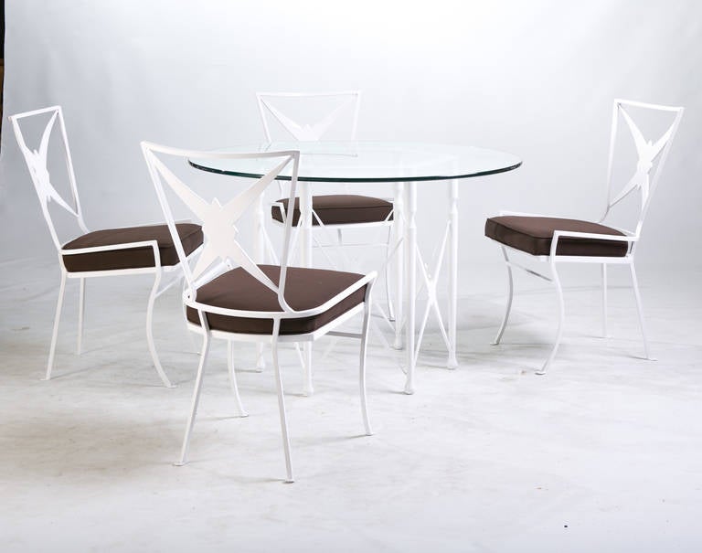 A Hollywood Regency dining set comprised of a round metal table and four chairs. Note the table's arrow motif base and the chairs' shield-like backs. The table and chairs have been newly powder coated bright white. The chair seats have been