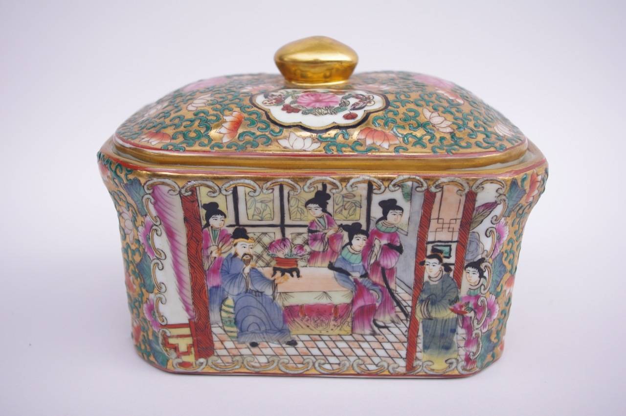 Covered rectangular box in Canton Chinese porcelain on a white background. Decoration with gold color with green foliage and stylized flowers. On one side, basket of flowers as dahlias and iris on white background, on the other side interior scene