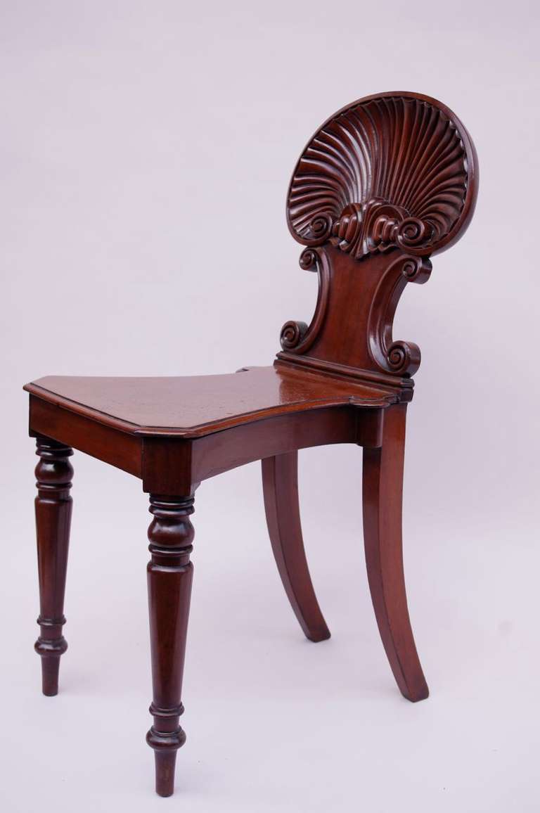 Regency style carved mahogany hall chair, standing on two turned tapered front legs and two saber back legs. Seat with canted corners tightened backwards. Back carved as a shell with two C-scrolls beneath.

Model created by Gillows of London and