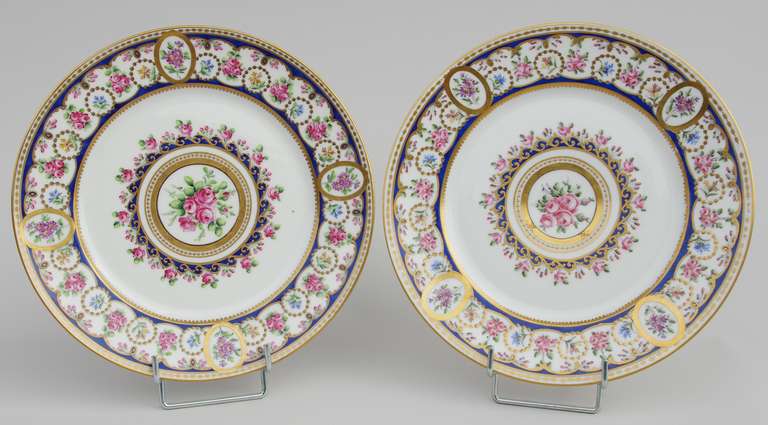 Pair of porcelain round plates with a white background and flowery decoration. Center is adorned with a bunch of roses circled with gilt nets and blue lapis waves. The wing is decorated with A same color banner on which a flowers frieze is deployed