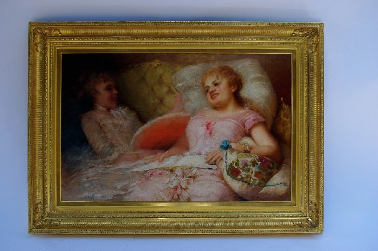 Framed oil on canvas representing two women in a couch. The right one is dressed in sweet pink and comfortably sat on wide pillows, a open book on her knees. Her friend is holding a red range and both of them are laughing, maybe because of their