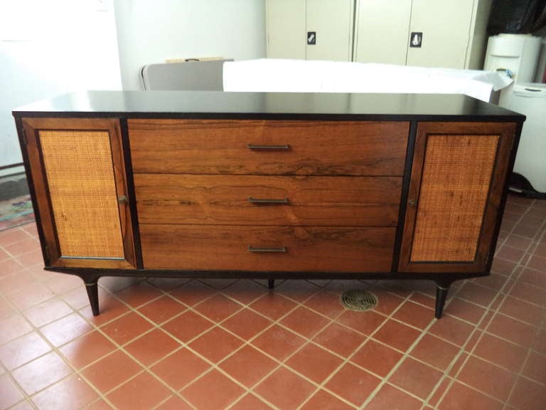 Mid Century Lane Credenza by Lane accentuated by a high gloss black finish and caning on the cabinet doors. There are also small brass trim pieces accentuating the pencil legs.This piece has three drawers down the middle, two cabinets on either side