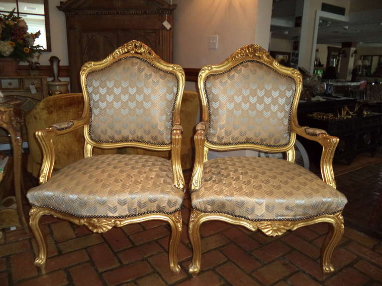 Pair of French Gilt fauteils in the style of Louis XVI Accentuated with open scroll arms, carved floral spray backs with serpentine seats  these elegant chairs are covered in a herringbone silk upholstery.