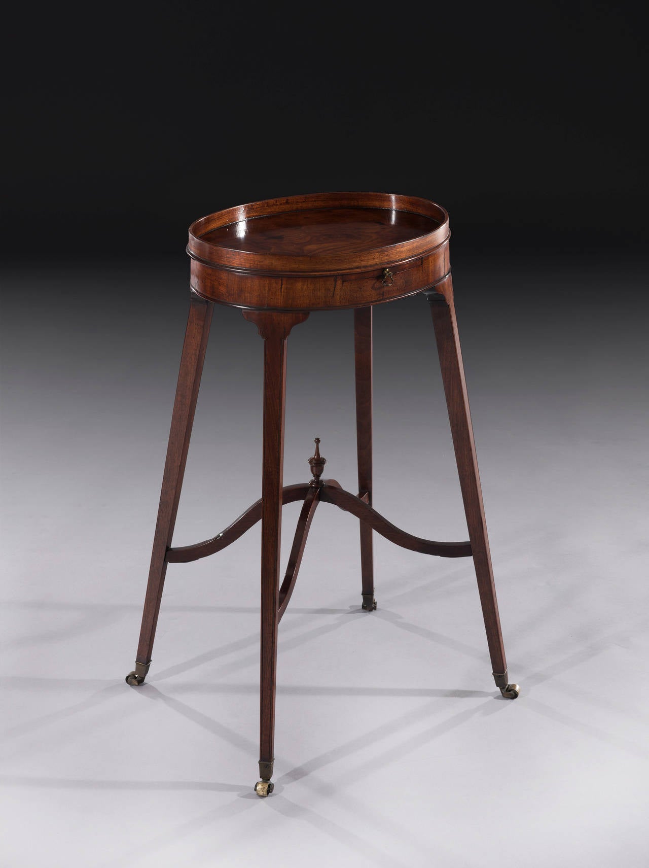 The George III oval shaped urn stand in the manner of George Hepplewhite retains a great colour and patina. The highly figured oval top with a low gallery sits on a mahogany veneered frieze with a centre pull-out slide. The four finely tapered legs