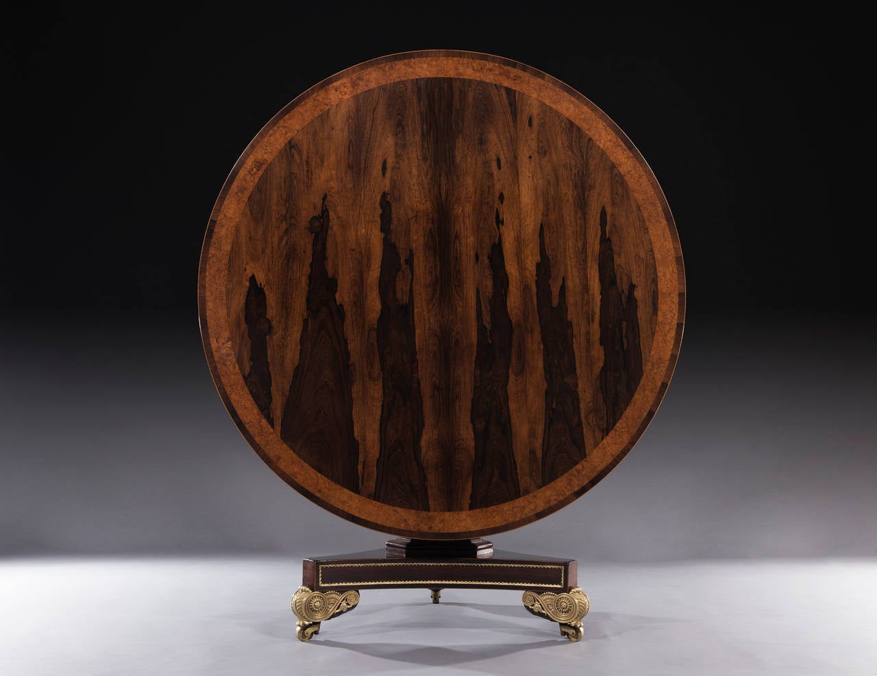 The Regency tilt top centre table with a circular rosewood veneered top, is cross-banded with a generous amboyna banding. The triform pedestal base with brass mouldings and inset rosewood panels is repeated on the plinth which sits on beautifully