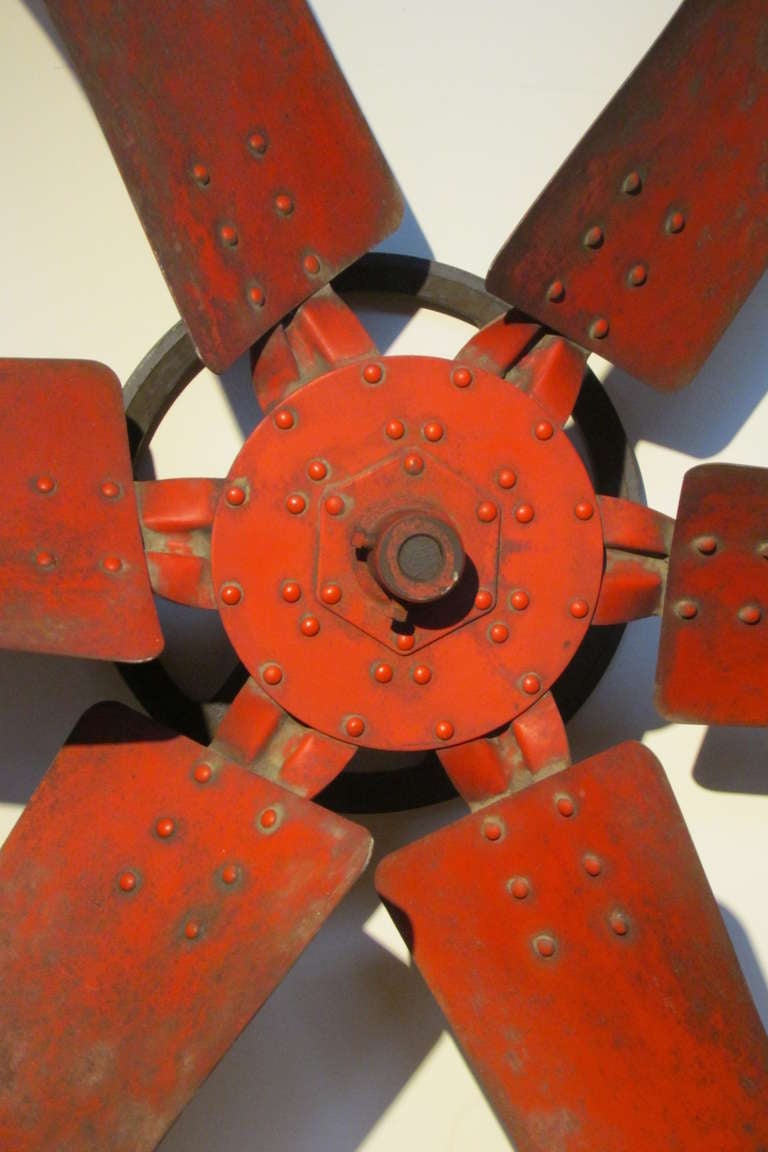 A large scale riveted steel fan blade with attached iron back wheel - once part of an industrial ventilation fan system -  brilliantly aged original persimmon red painted surface with just the right amount of wear - an edgy sculptural object that