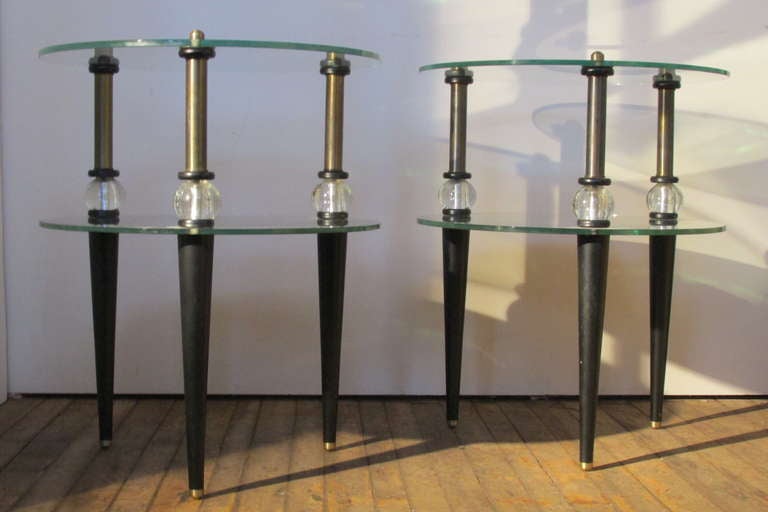 Dating from the the mid 20th century a glamorous pair of  thick beveled plate glass & mirrored two tiered tables in the Art Deco style of Gilbert Rohde  - each with three tapered black ebonized wood legs & upper bronzed brass posts mounted with