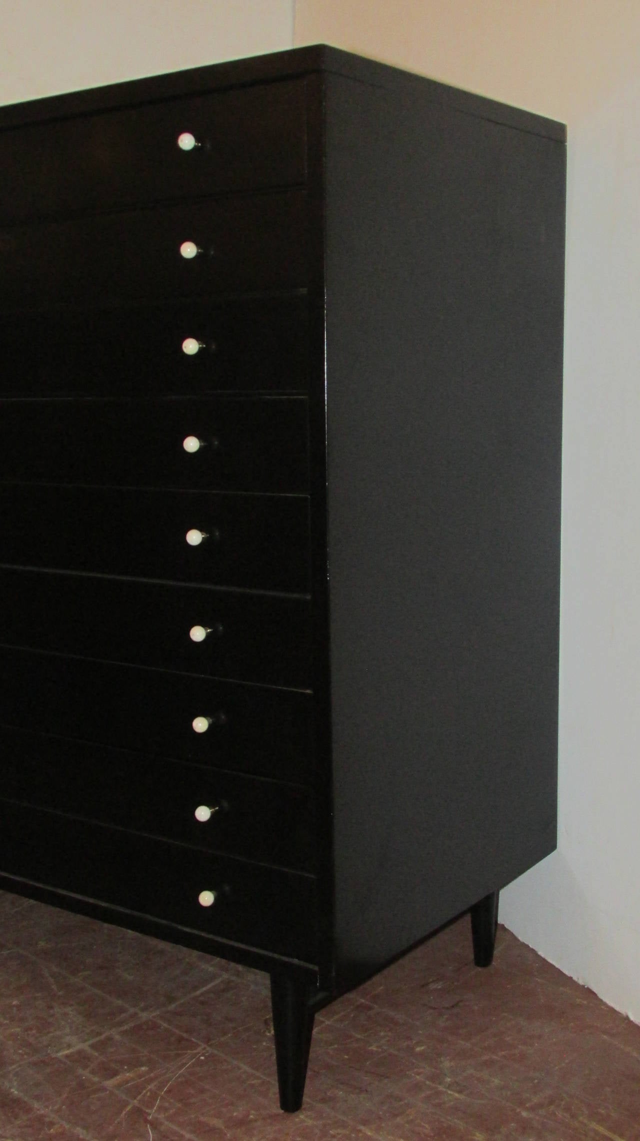 In the modernist style of Paul McCobb, a five-drawer tall chest highboy in beautiful black ebonized lacquered surface raised on four tapered legs joined by side stretchers. Retains the original white laminate top and pearly white ceramic ball and