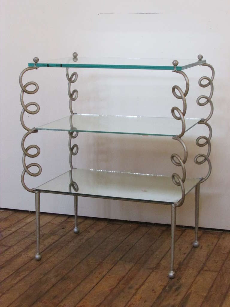 A silver painted hand wrought spiral metal table with four lower ball feet and four upper ball finials that go through circular holes in the top beveled plate glass shelf and screw into the framework of the table. The two lower shelves are set with