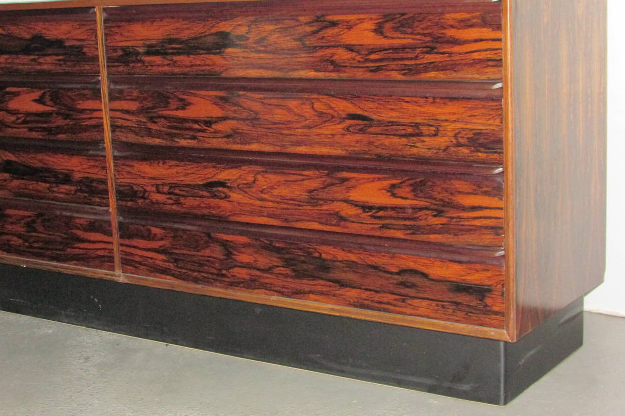 A Scandinavian modern eight-drawer long chest, credenza, sideboard in vibrantly figured book matched Brazilian rosewood veneer on front, sides, top, finished back - the recessed base with a nicely contrasting black rubberized molding on all four
