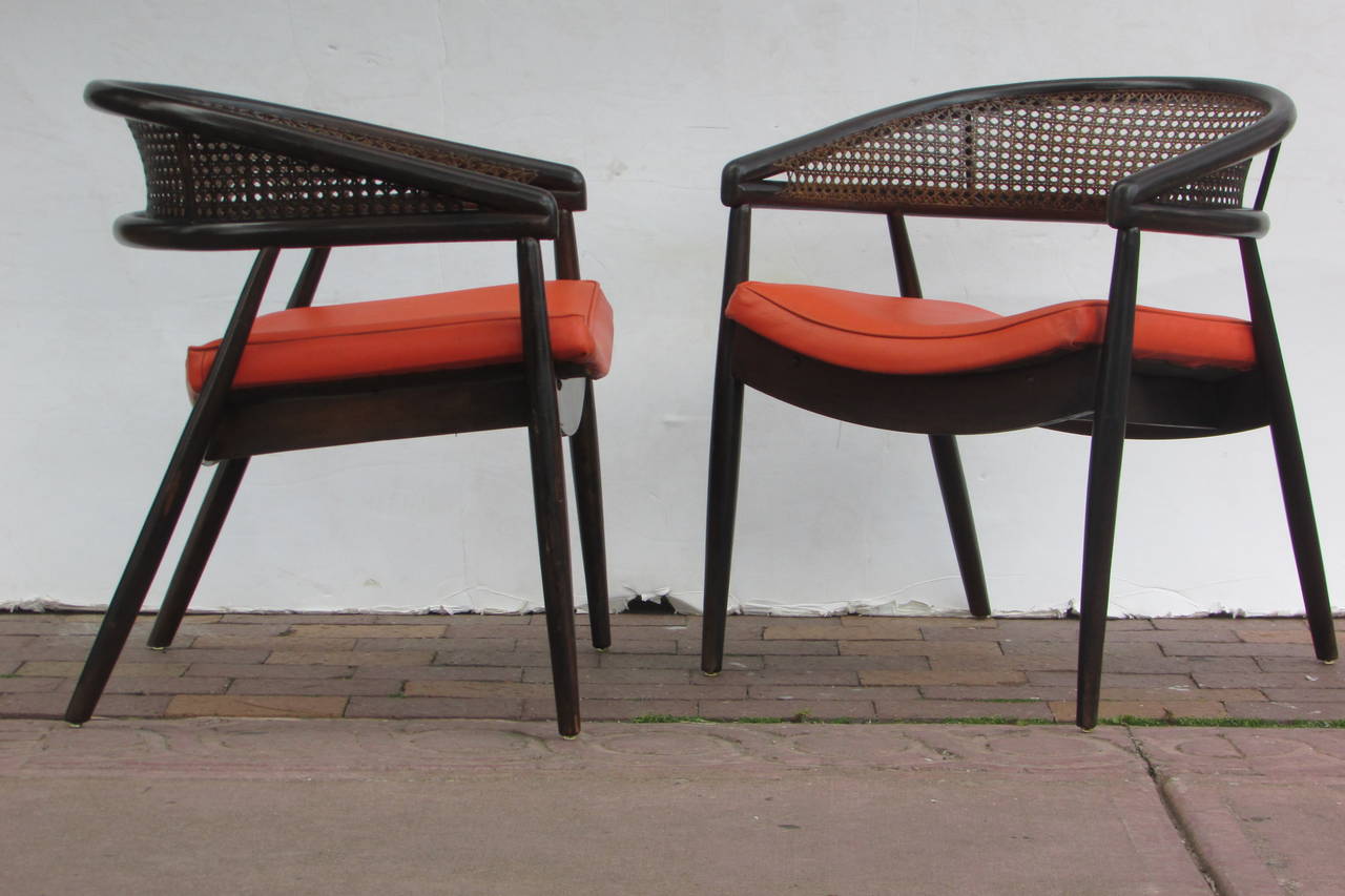 Pair of caned rounded back lounge chairs with angled sculptural form in all original dark brown to black stained finish and orange vinyl upholstered seats. These same chairs were used by James Mont for his design commission of the King Cole