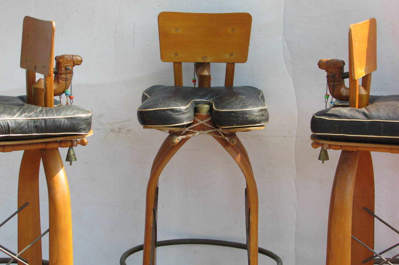 Three funky decorative 1960s Moroccan bar height carved camel stools with original black leather seat pads / brass posts under seats / brass tacking on legs / metal x stretchers / brass metal ring foot rests / rawhide / hanging tassels /  beads and