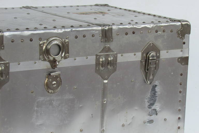 A large industrial riveted aluminum steamer trunk with with old paper labels on exterior that read 