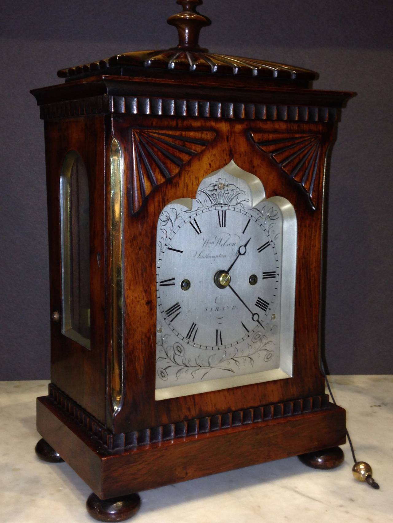 A very small attractive rosewood library table clock by William Wilson Southampton Street. Strand London from 1839 -44. The case has a gadrooned top with a turned finial, canted side angles with brass inlay, and a base with a decorative moulding