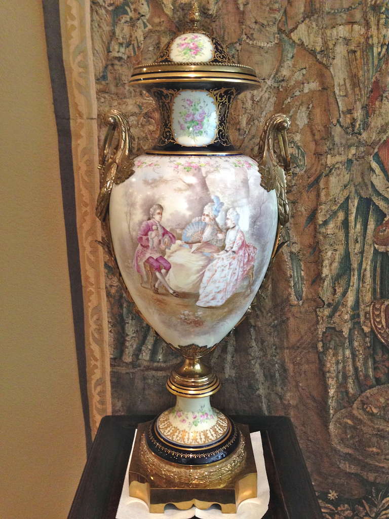 A beautiful hand-painted interior scene decorates this palace urn painted in the round, the reverse a peaceful landscape is the subject. Gorgeous raised paste gilt is highlighting cartouches containing delicate paintings of pink roses.