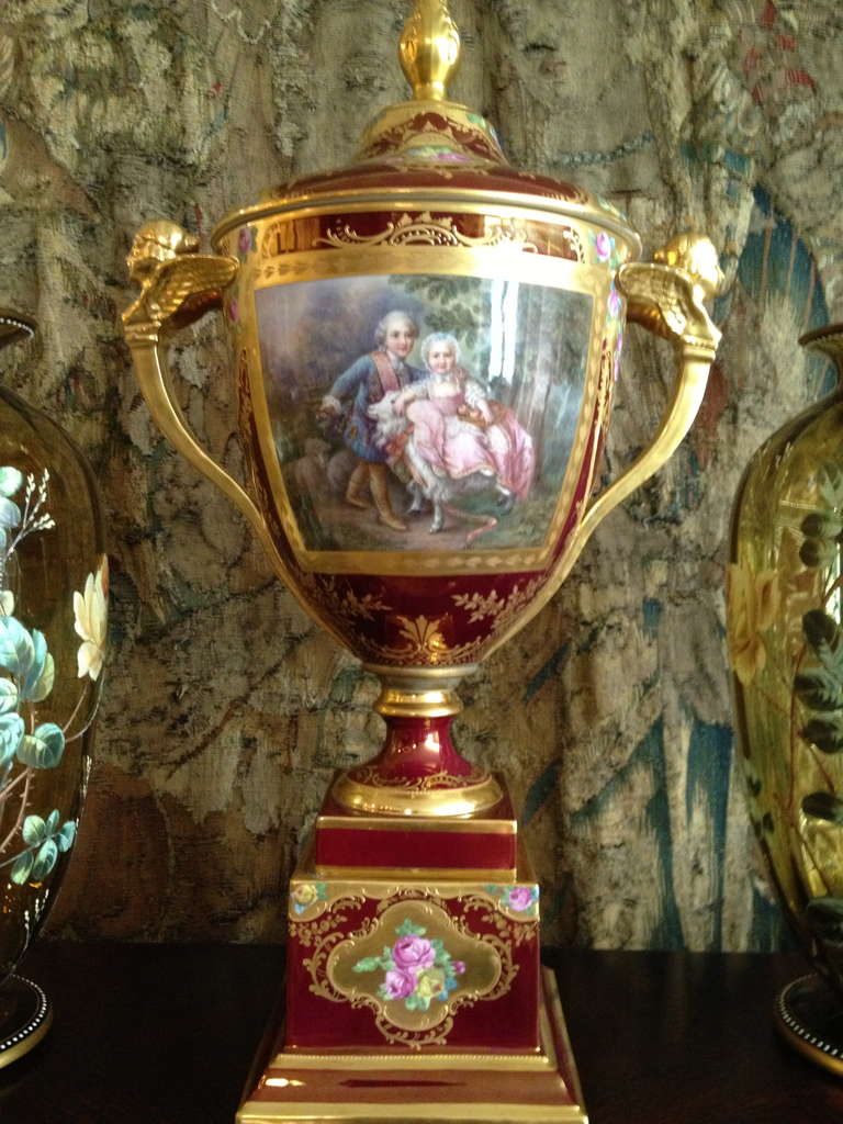 This lovely Royal Vienna style vase has a beautiful rich red iridescent ground glaze color. The hand painted scene with 
a charming painting depicting children all dressed up like adults
In 18th century attire. Gilt cartouches containing