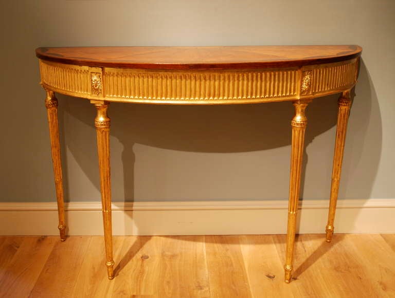 A late 18th Century demi lune side table, the top veneered in satinwood, crossbanded and inlaid with neo-classical motifs. The carved and gilded base with flutted frieze and finely tapering flutted legs. Circa 1780.