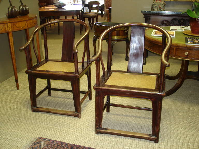 A pair of Chinese horseshoe back chairs. Of soft wood, with a red, possibly cinnabar glaze, the arching three part back supported by curving backsplats, with mitred square frames supporting the woven rush seats all supported by slightly splayed legs