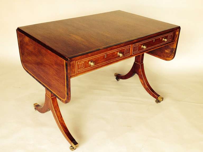 This table is a classic Sheraton period sofa table with elegant swept end supports and brass cap casters. It is an amazing golden colour and has contrasting rosewood cross banding around the top edge and box wood line inlay throughout.
