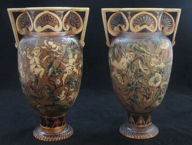 Pair of Martin Brothers Two Handled Vases decorated with Cherubs amongst flowers and foliage. Hairline crack to interior of one neck (doesn't appear to travel to exterior)