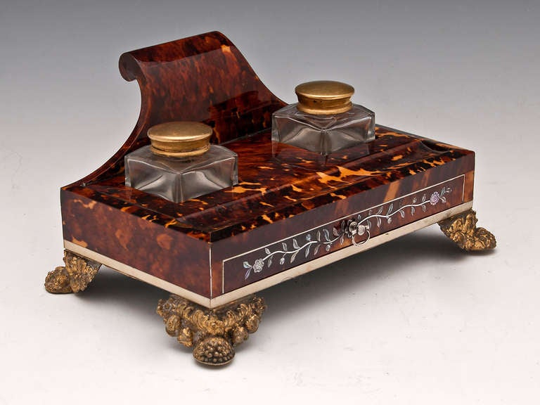 This impressive regency tortoiseshell ink stand is inlaid with Mother of Pearl flowers and silver stringing, it stands on four gilded pressed brass lion pad feet and has tow original glass inkwells with gilded screw top lids. 

The bottom of this