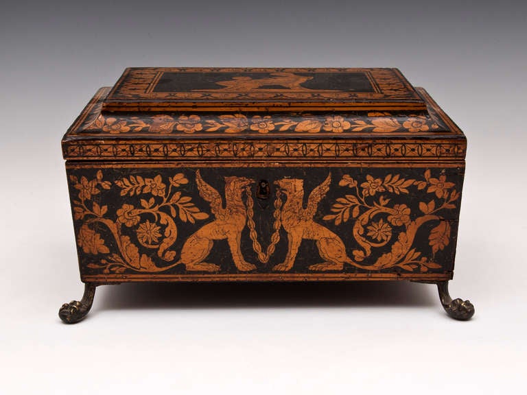 Penwork sewing box with scrolling leaf design, Griffins on the front and back and a lovely picture of a mother and child on the top. The sides feature brass lions head swing handles, while the box itself stands on four brass lions feet. 

The