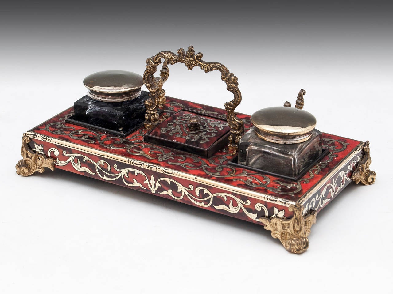 Boulle Inkstand with decorative gilded Ormolo brass feet, pen holder and carrying handle. To the centre is a small Boulle lidded compartment, either side of this it has two brass screw top inkwells. 

The 
