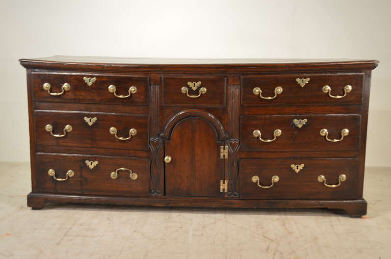 18th century English oak dresser base with a three plank top with a beveled edge. The case is comprised of two banks of three drawers each flanking a central single drawer over a paneled door. Raised on bracket feet.