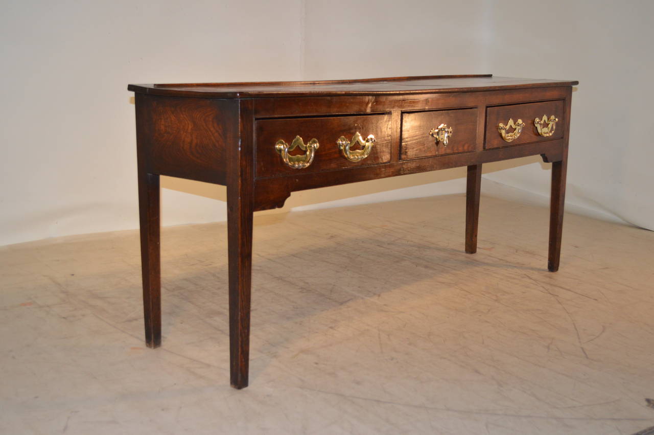 18th century English sideboard made from elm. The top is made up of one single plank with a plate rail attached on the back. The apron contains three drawers with fabulous oversized brass hardware, and follow down to nicely tapered legs.