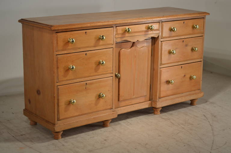 19th century English dresser base made from pine. The top is made up of two planks, and they have a beveled edge following down to a central drawer over a central door, which opens to reveal two shelves. These are flanked by two sets of three