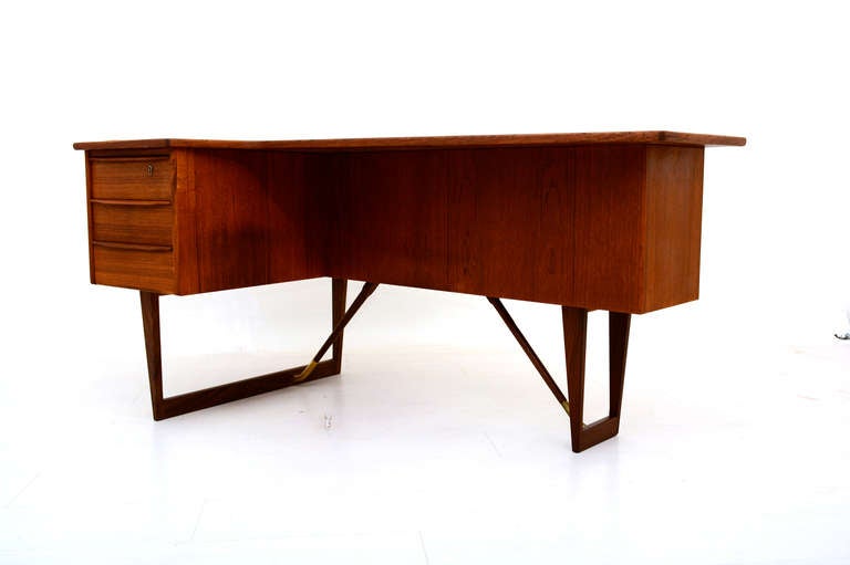 Teak Desk by Loving, made in Denmark.

Receiving desk with unusual boomerang shape. Mounted in sculptural legs with brass fittings.

The receiving part has an open storage and closed compartment for a small dry bar. 


