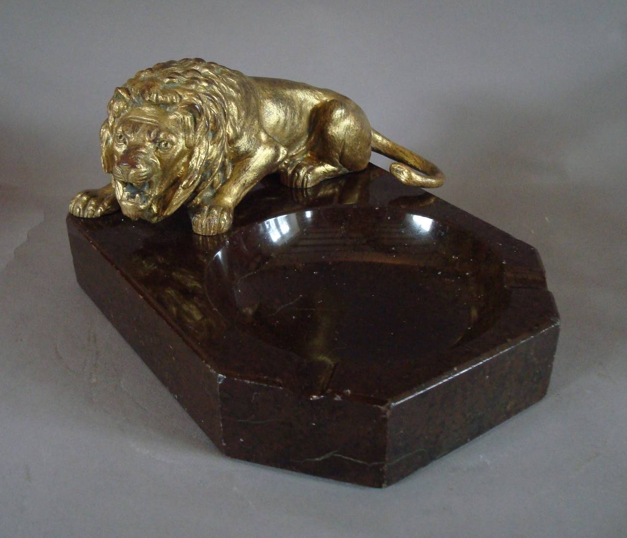 Early C20th lion and marble ashtray, the gilt bronze crouching lion standing on the substantial 'Serpentine' marble ashtray base incorporating a circular ash bowl with a cigar/cigarette rest to each canted corner.

Also suitable to use as a desk