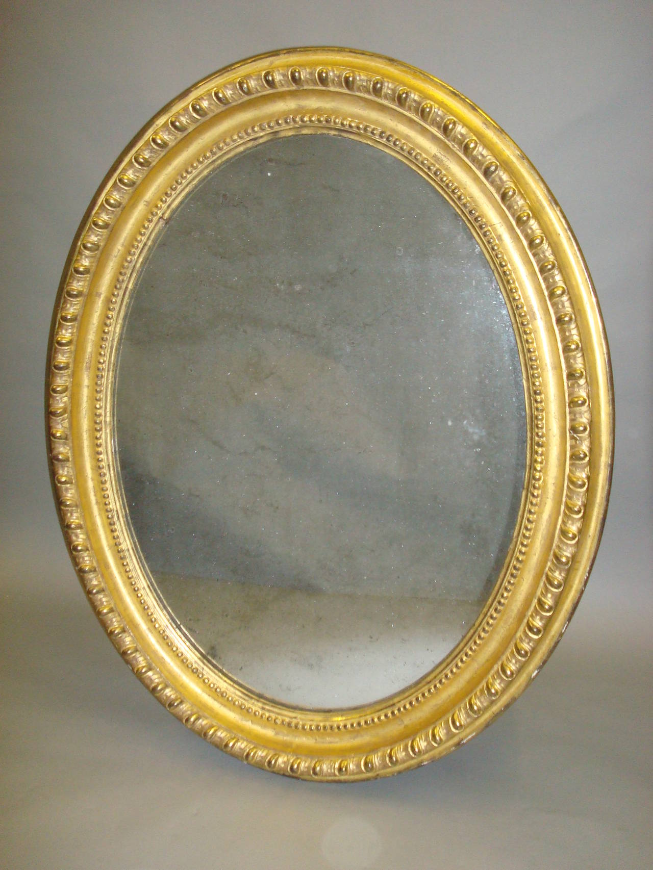 An early C19th giltwood oval wall mirror; the bold shaped frame with a prominent egg and dart border and a smaller beaded inner moulding, housing the original mercury mirror plate which has achieved a small amount of distressing.