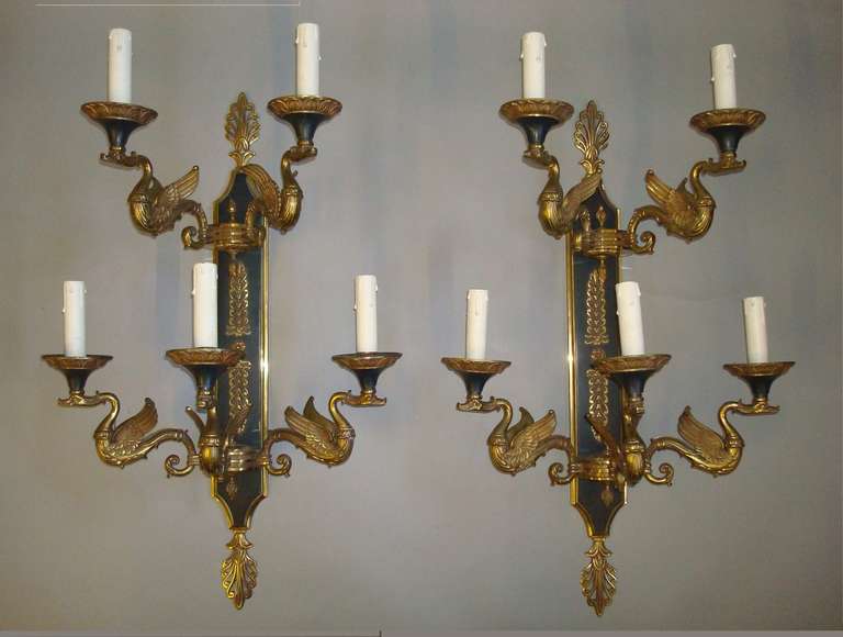 An exceptional Empire style set of four gilt brass wall sconces/lights, the rectangular backplate with dark green patinated finish and stylised decoration with anthemion cresting and terminals, supporting two circular roundels with flame finials. 