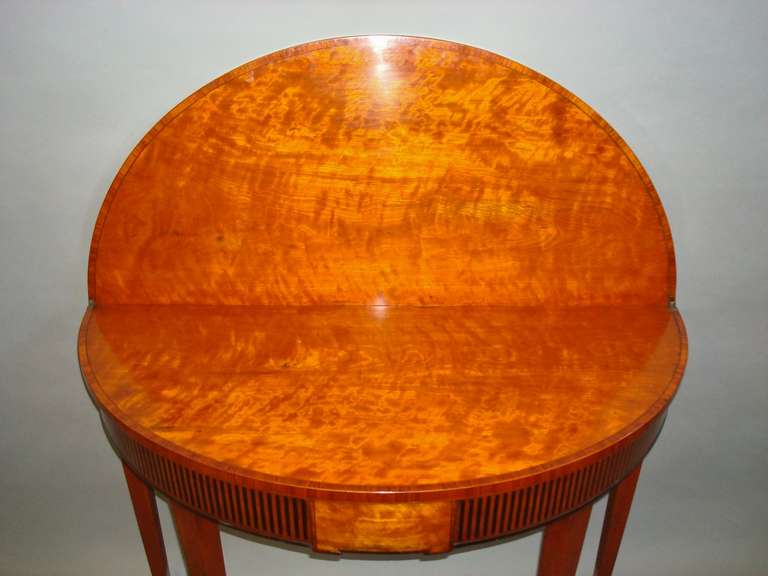 A good George III satinwood semi eliptical fold over tea table; the shaped top with tulip wood cross banding opening to reveal an oval shaped well figured interior also cross banded with tulip wood; raised on a frieze with ebony inlay with central