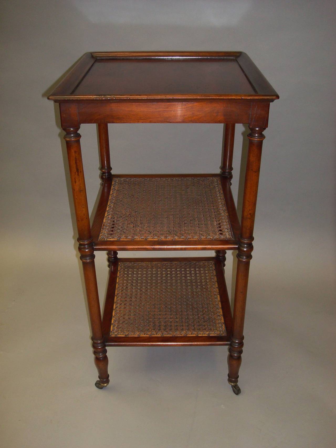Regency mahogany three tier étagère, the figured mahogany top with a shaped raised moulding to the sides, on slender turned supports, united by two lower tiers with cane inset panels, raised on turned tapering legs with original brass castors.
