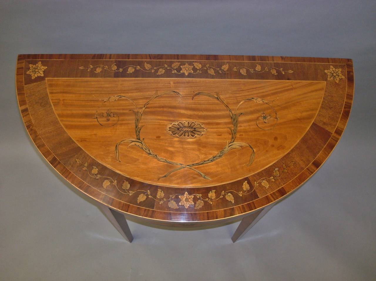 A fine George III satinwood and marquetry demilune card table; the half moon top with a large satinwood panel centred with an oval sycamore paterae surrounded by scrolling foliage; with a wide border having large flower heads with trailing leaves