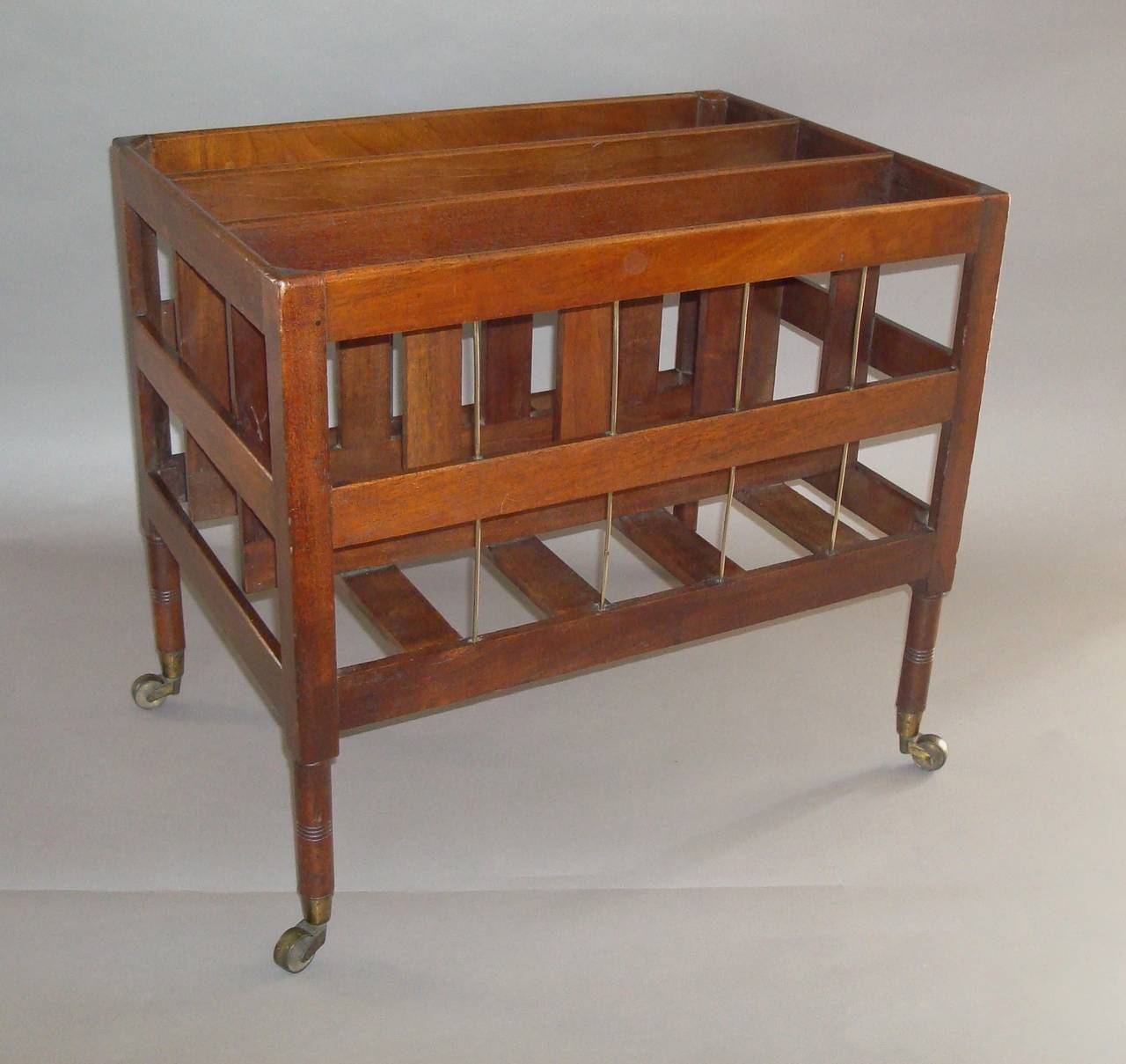 An elegant Regency mahogany canterbury of unusual open slatted form; with three interior divisions, the front and reverse incorporating two rows of vertical brass rods, raised on slender turned legs with original brass cup castors.