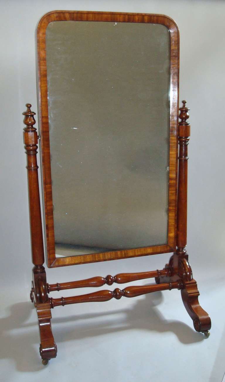 A good quality George IV mahogany cheval mirror; the rectangular mirror plate enclosed in a figured mahogany frame, supported on turned tapering columns with urn shaped finials, raised on scrolled legs with roundels to either side united by twin