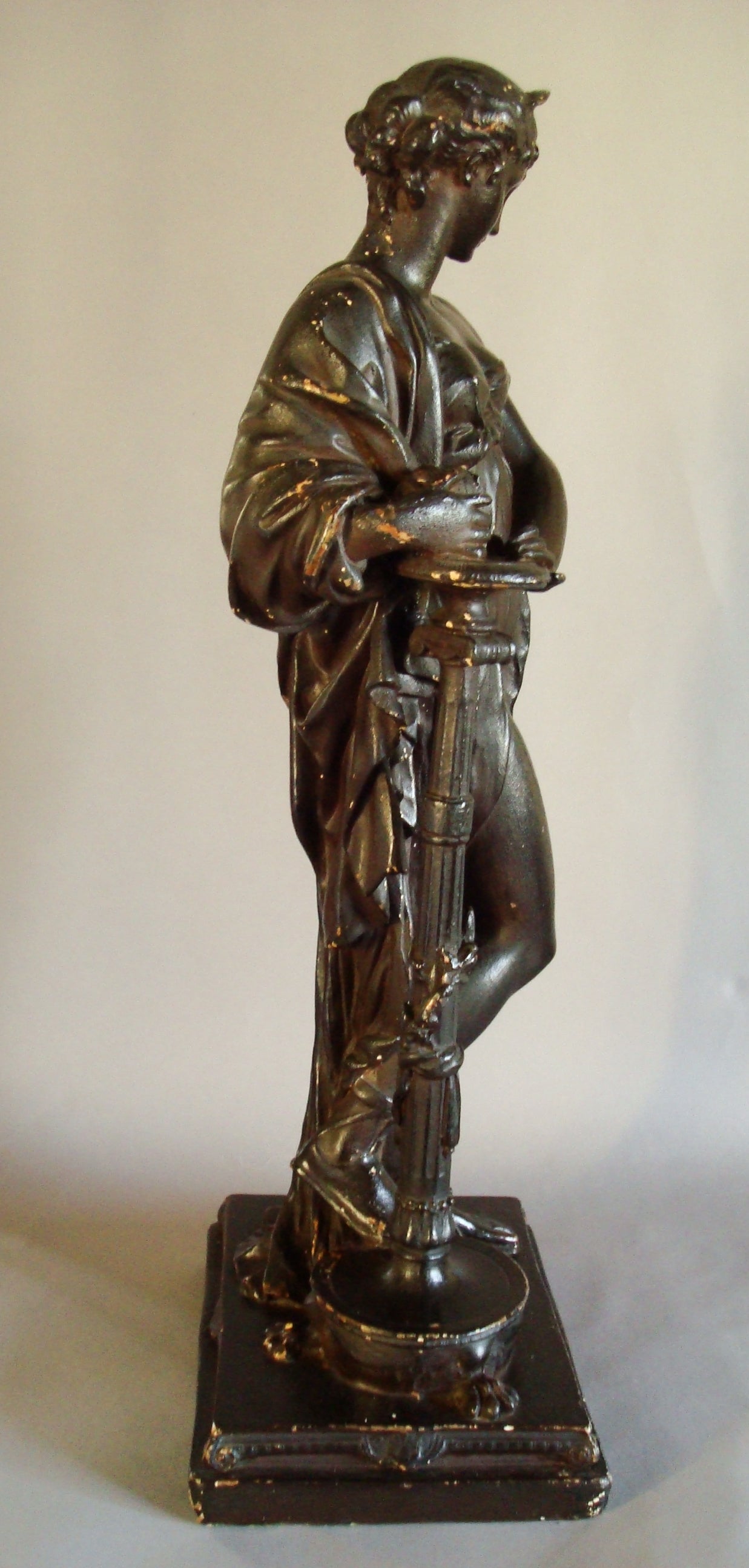 A Regency 'bronzed' plaster figure of a classical lady; the semi-clad figure with tied back hair and head dress, holding a vessel under her arm, resting on a Thomas Hope inspired classical torchere with paw feet.  Her right leg raised on the