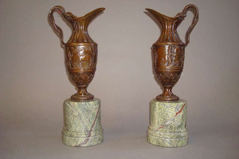 C19th French pair of bronze ewers, the fluted neck with acanthus leaf and entwined snake handle above a freize with an interesting scene of figures and horses, the 'waisted' base with acanthus leaf decoration standing on a plinth of olive green