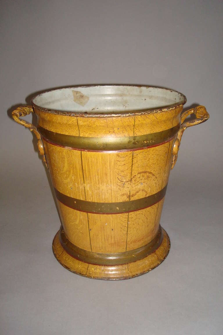 Mid 19th century very unusual japanned tole bucket/jardinere/wine cooler; of circular tapering form; the side carrying handles scrolled with acanthus leaves; the original japanned decoration simulating pale oak with brass bands.  Marked Shoolbred