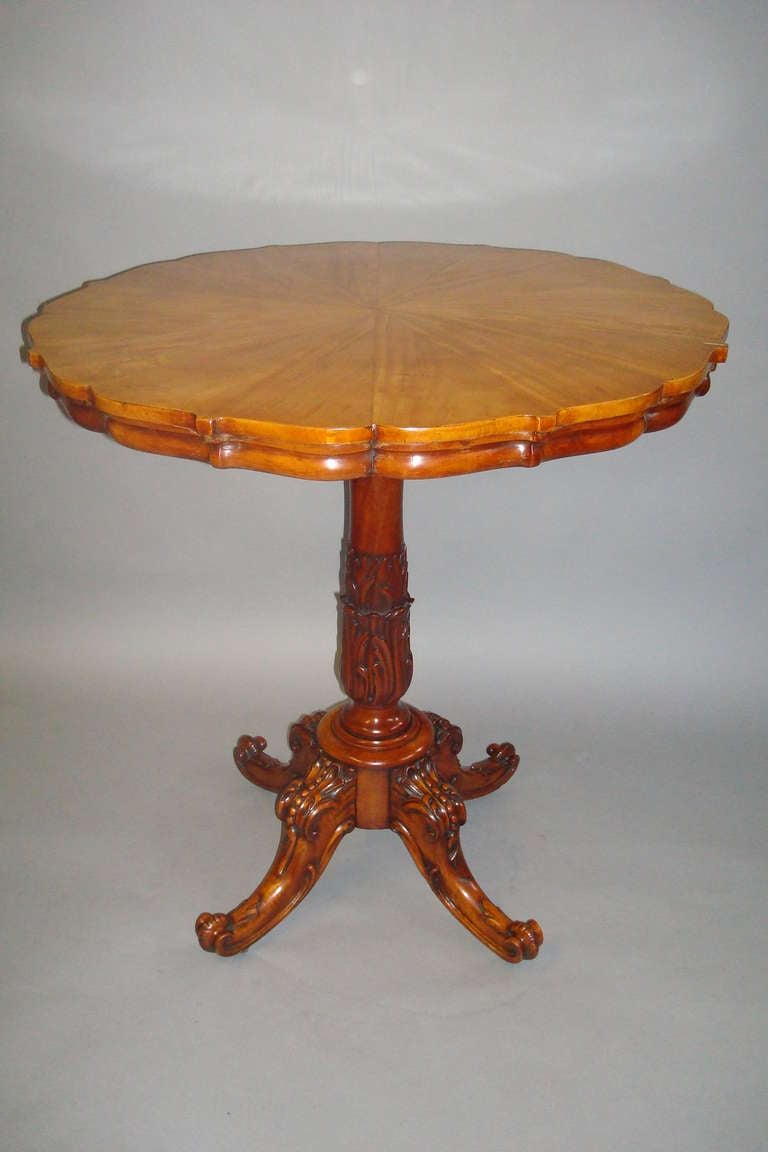 A good quality George IV satinwood occasional/centre table, the circular top with a serrated edge, veneered in radiating figured satinwood; with a shallow moulded frieze below; raised on a central column in solid satinwood with carved palm leaves;