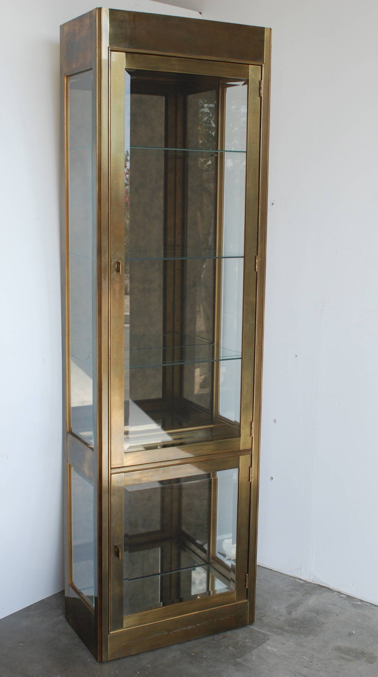 A lighted antique brass vitrine made by Mastercraft. There are two doors fitted with beveled glass. The top section has three adjustable shelves. There is smoked antique mirrored backing and a mirror bottom. 
The 14 3/4