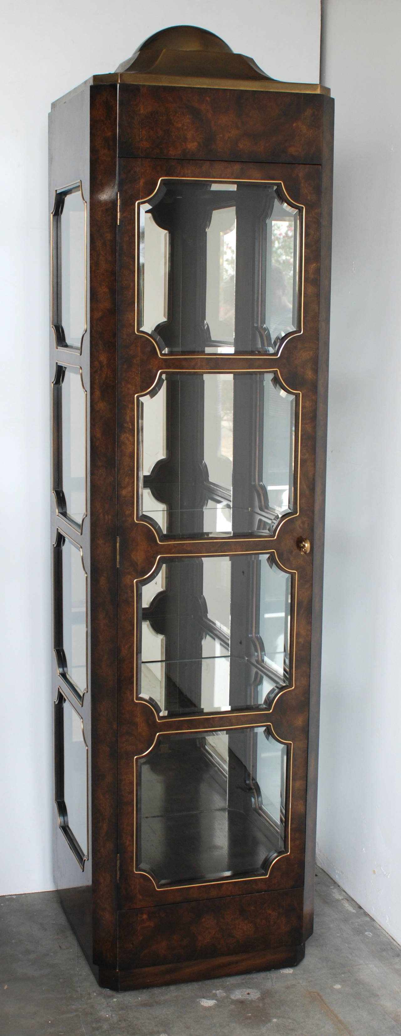 A beautiful high quality vitrine made of highly figured rich dark burl wood and capped with a brass dome. The cabinet lights up and also retains its original pull. It has three glass shelves and a wooden base platform. The geometric glass