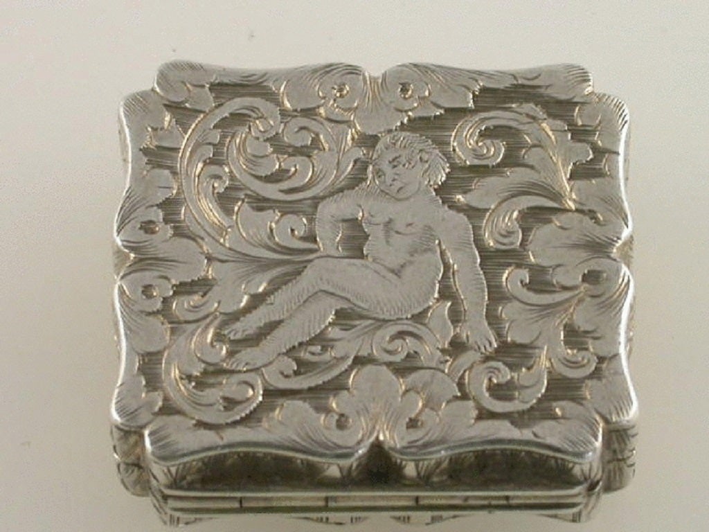 An exquisite early Victorian small silver Vinaigrette of shaped rectangular form, the base very finely engraved with a reclining naked cherub amongst foliate scroll work, the sides with geometric designs, the lid with a central cartouche engraved
