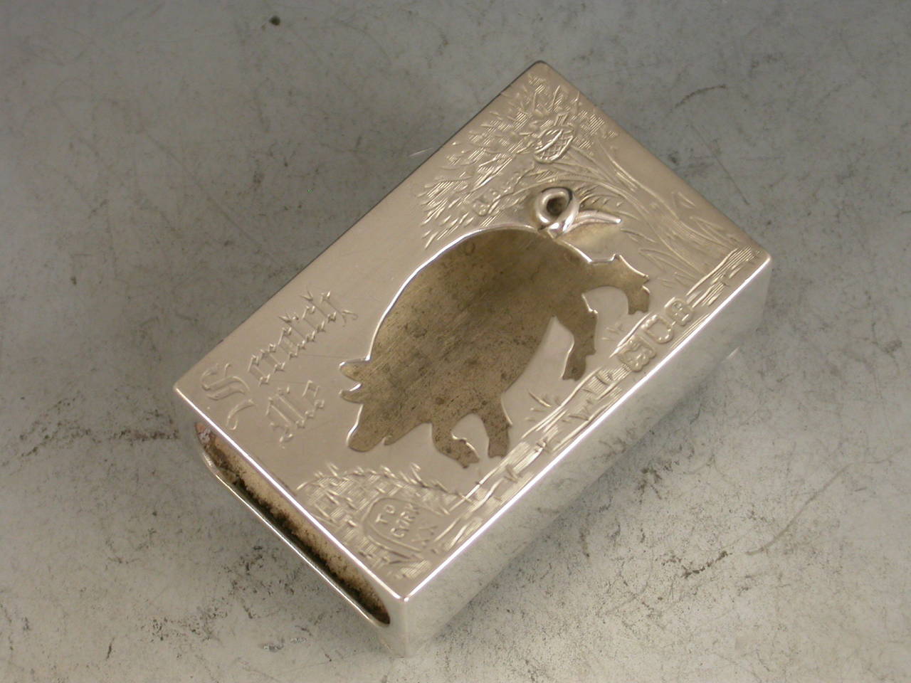 A comical Edwardian novelty silver Matchbox Cover standing on four ball feet, the cover with cut out silhouette of a pig going to Cork and engraved with the legend "Scratch Me". A bird sitting on a nest in a tree to the right hand side.