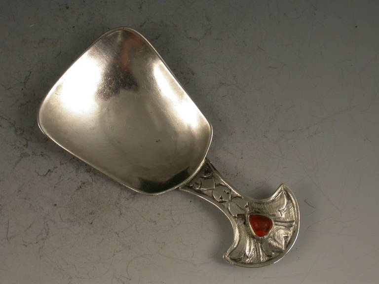 A stylish early 20th century silver Caddy Spoon made in the Arts & Crafts style with a scoop shaped bowl and a chased handle with fan shaped terminal set with an amber coloured heart shaped stone, the reverse of the handle inscribed; 