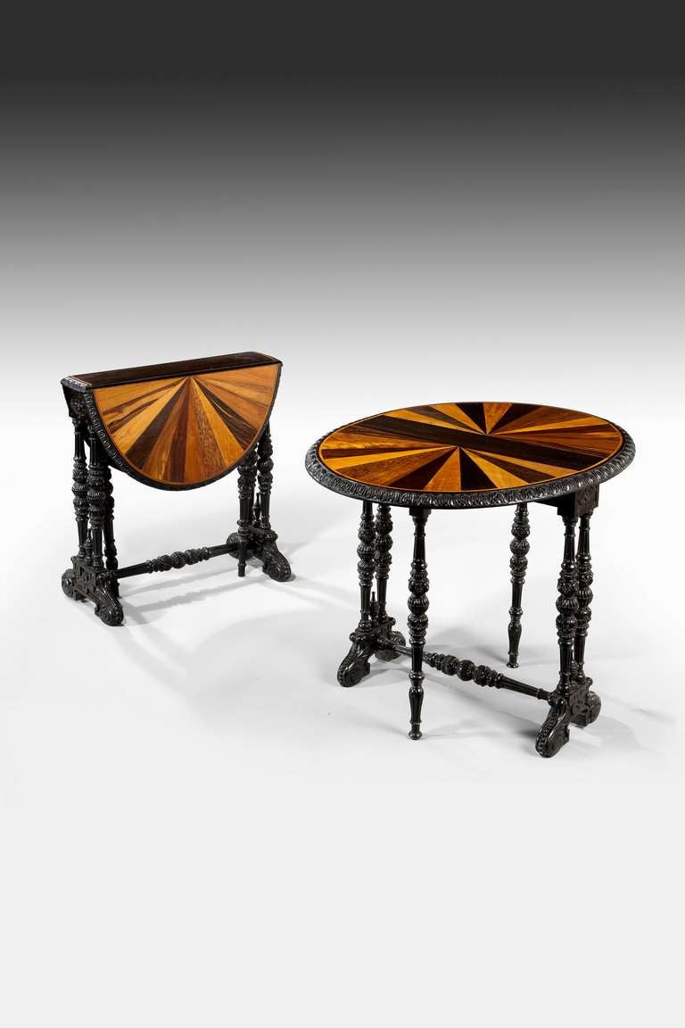 These matched Colonial tables are unusual in their construction. They are of typical oval form with carved and turned ebony gate legs and fall flaps on either side. The decoration consists of a central ebony panel flanked by radiating segments of
