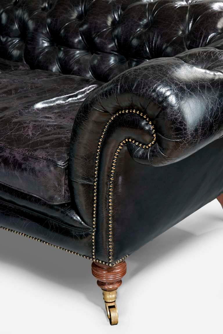 An antique black leather Chesterfield by Howard and Sons 
This sofa has a deep-buttoned roll over back with scrolling arms set upon turned spinning top front feet. The splayed back legs stamped with a number.